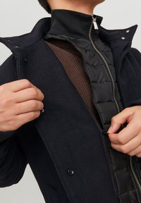 Abrigo de mezcla de lana azul marino con un forro acolchado negro, cierre con cremallera y cuello alzado. Manos ajustan la parte delantera del abrigo.