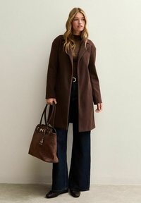 Cappotto in misto lana marrone con vestibilità sartoriale, abbinato a jeans a zampa d'elefante scuri e una borsa a tracolla in suede marrone. Presenta una cintura e calzature eleganti.