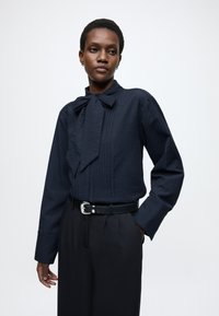 Tmavomodrá textúrovaná blúzka s mašľou na golieri, dlhé rukávy a vzor s vertikálnymi pruhmi, skombinovaná s čiernymi nohavicami s vysokým pásom.