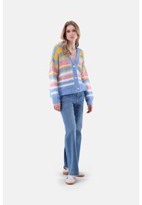 Multifarbene Strickjacke mit tiefem V-Ausschnitt, ausgestattet mit Streifen in Blau, Gelb, Pink und Weiß, kombiniert mit hellblauen Jeans und weißen Sneakern.