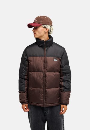 Junger Mann trägt eine braune und schwarze Steppjacke und eine braune Kappe, blickt nach links vor einem einfarbigen hellen Hintergrund.