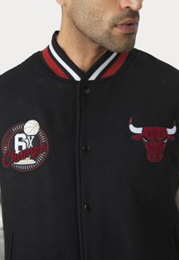 Melnā vilnas varsity jaka ar sarkanbaltiem svītrām apkaklē, izšūtu sarkanu bullīša logotipu un apļveida uzšuvumu ar uzrakstu "6X čempioni".