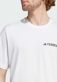 Bílý bavlněný tričko s krátkým rukávem, kulatým výstřihem a černým logem "TERREX" na levé části hrudi. Hladká textura, volnější střih.