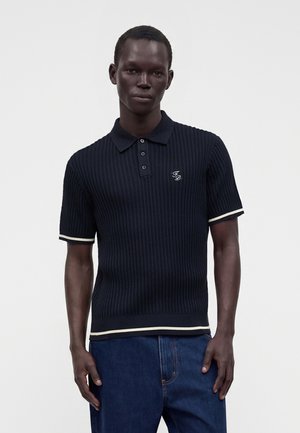 Ung mand iført en sort ribbet kortærmet poloshirt med hvide kanter og blå jeans, stående foran en ensfarvet baggrund.