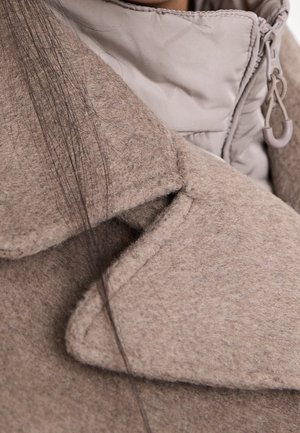 Close-up van een beige wollen jas kraag, gelaagd over een lichtgrijze gewatteerde jas met zichtbare ritstrekker en lange, donkere haarlokken.