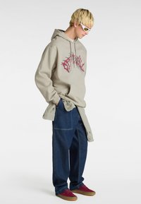 Beige hoodie met rode grafische ontwerp, gelaagd over een lichtgrijze shirt, gecombineerd met losse donkere spijkerbroeken en bordeauxrode skate schoenen.