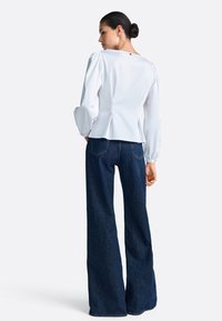 Biela blúzka s dlhým rukávom, naberanými rukávmi a priliehavým pásom, v kombinácii s tmavomodrými džínsami so širokými nohavicami, zadnými vreckami a neupraveným lemom.