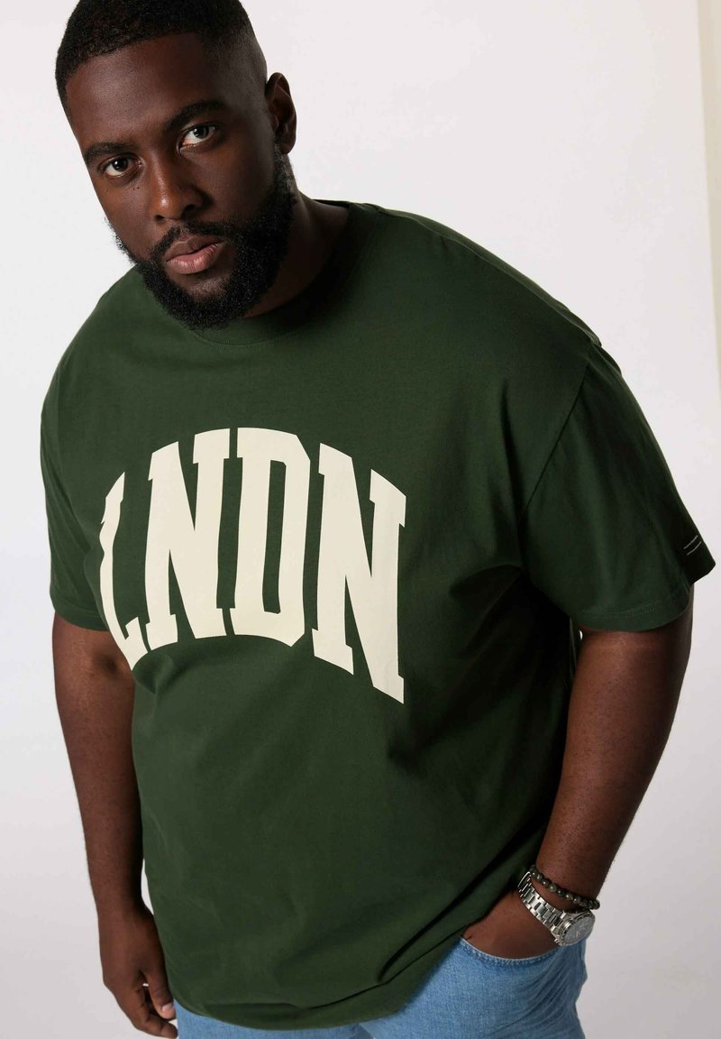 Mørkegrønne bomulds t-shirt med "LDN" i cremegul fed skrift. Har korte ærmer og en afslappet pasform. Bæres med lyseblå jeans.