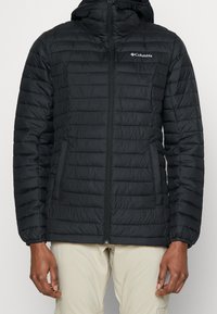 Giacca invernale nera isolata con zip frontale, polsini elasticizzati e cappuccio. Presenta un design a quadri orizzontali e il logo Columbia sul petto.