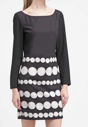 Femme portant une robe noire à manches longues avec des rangées horizontales de grands et petits cercles blancs sur la jupe et la zone de la taille.