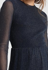 Robe bleu marine avec de longues manches transparentes, en tissu texturé avec des paillettes dorées, taille froncée et encolure ronde.