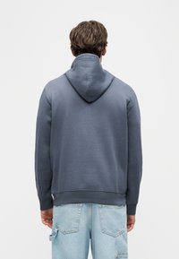 Άνδρας που φοράει σκουρόχρωμο γκρι φούτερ με κουκούλα και ανοιχτό μπλε τζιν με πίσω τσέπες, γυρισμένος με την πλάτη σε απλό λευκό φόντο.