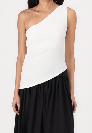 Kobieta ubrana w biały, asymetryczny top bez rękawów połączony z czarną, marszczoną spódnicą z wysokim stanem na jednolitym tle.