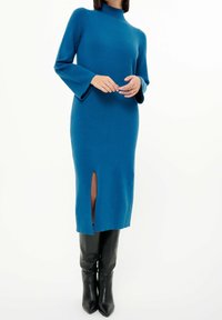 Stickad teal midiklänning med hög krage, utsvängda ärmar och slits fram, i kombination med knähöga svarta läderstövlar.