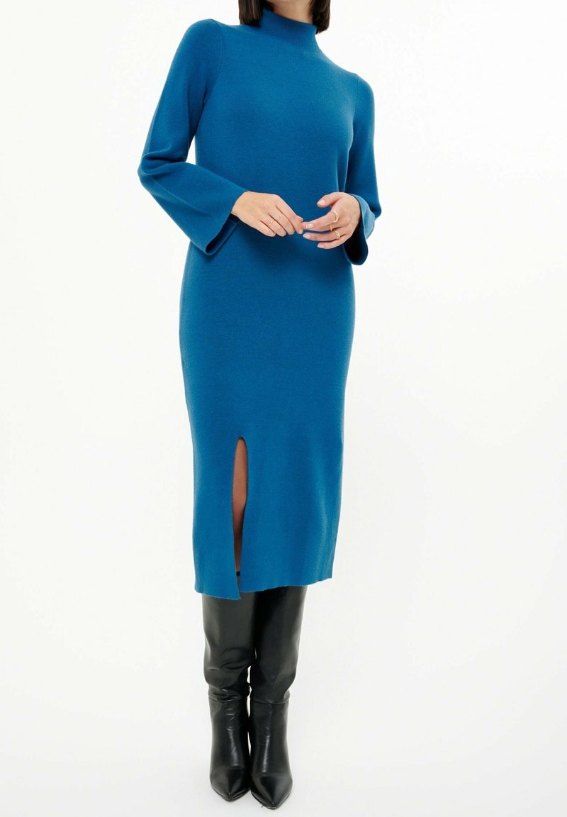 Stickad teal midiklänning med hög krage, utsvängda ärmar och slits fram, i kombination med knähöga svarta läderstövlar.