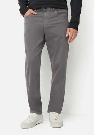 Mann trägt graue Freizeithose, weiße Sneakers und ein schwarzes Langarmshirt, steht mit einer Hand in der Tasche.