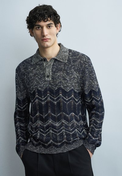 Jeune homme aux cheveux courts et bouclés portant un pull en maille à motif chevrons foncé et un pantalon noir, debout devant un fond clair uni.