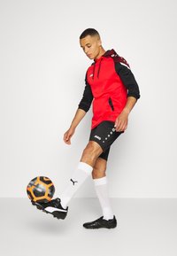 Roter und schwarzer Sporthoodie mit Kapuze und Zugband, kombiniert mit schwarzen Shorts mit weißen Logoakzenten, weißen Socken und schwarzen Fußballschuhen.