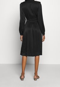 Musta mekko pitkillä hihoilla, kyynerpäähän asti ylettyvällä leikkauksella ja vyötäröltä kiristettynä; siinä on hienovarainen chevron-kuvio ja sileä rakenne.