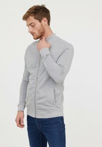 Veste grise zippée en tissu doux, avec un col montant et des poches latérales, portée avec un jean bleu. Design sans coutures avec des poignets côtelés.