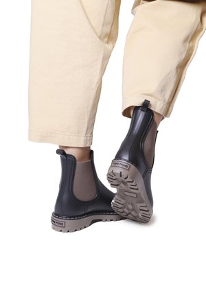 Bottines Chelsea noires avec des panneaux latéraux élastiques texturés et une semelle en caoutchouc beige. Comprend une languette et un marquage sur le talon.