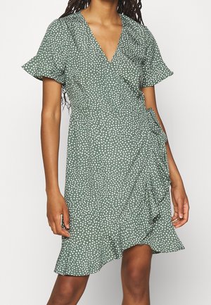 Vestido cruzado verde con lunares blancos, mangas cortas con volantes y dobladillo fruncido, llevado por una persona de pie.