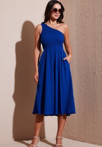 One-shoulder blue dress made of textured fabric, fitted bodice, flared skirt with pockets. Worn with sunglasses and strappy sandals.