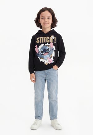 Enfant debout portant un sweat à capuche noir avec le dessin animé Stitch et un jean bleu clair, baskets blanches, main dans la poche, souriant sur fond blanc.