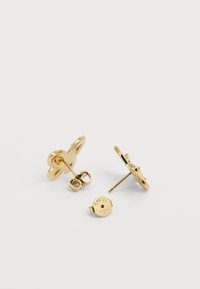 Boucles d'oreilles en or en forme de fleur, avec une finition lisse, une petite pierre précieuse transparente en accent, et un dos marqué du nom "Guess."