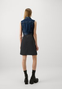 Gilet in denim, gonna grigia scura, silhouette dritta, design senza maniche, vita regolabile, indossato con stivali neri con tacco. Texture liscia.