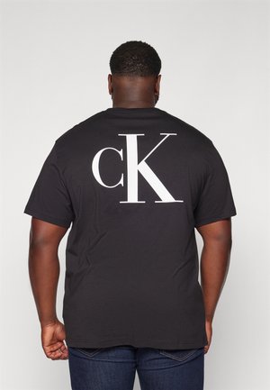 Mann trägt schwarzes Kurzarm-T-Shirt mit großem weißem "cK" Logo auf dem Rücken, kombiniert mit dunkelblauen Jeans, mit dem Rücken zur Kamera.