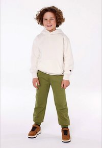 Χαρούμενο παιδί με σγουρά μαλλιά, ντυμένο με λευκή φούτερ, ελαιόχρωμα παντελόνια και καφέ αθλητικά παπούτσια, στέκεται μπροστά από λευκό φόντο.