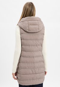 Hellbeige wattierte Weste mit Kapuze, die ein gesteppter Muster und eine glatte Textur aufweist. Getragen über einem cremefarbenen Strickpullover.