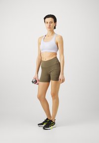Lys lilla sportsbh og olivengrønne højtaljede shorts, parret med sorte sneakers med neonlys. Holder en lyserød vandflaske.
