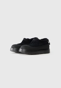 Black slip-on shoes with thick rubber soles and textured toe caps, featuring soft fabric uppers and a pull tab at the heel.