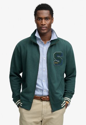 Mężczyzna noszący ciemnozieloną kurtkę na suwak z dużą literą „S”, niebieską koszulę w paski, beżowe spodnie, pleciony pasek i zegarek, ręce w kieszeniach.