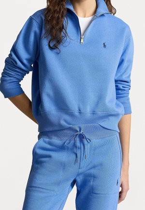 Personne portant un sweat-shirt bleu clair à fermeture éclair quart et un pantalon à cordon assorti avec un petit logo brodé sur la poitrine gauche et la cuisse.