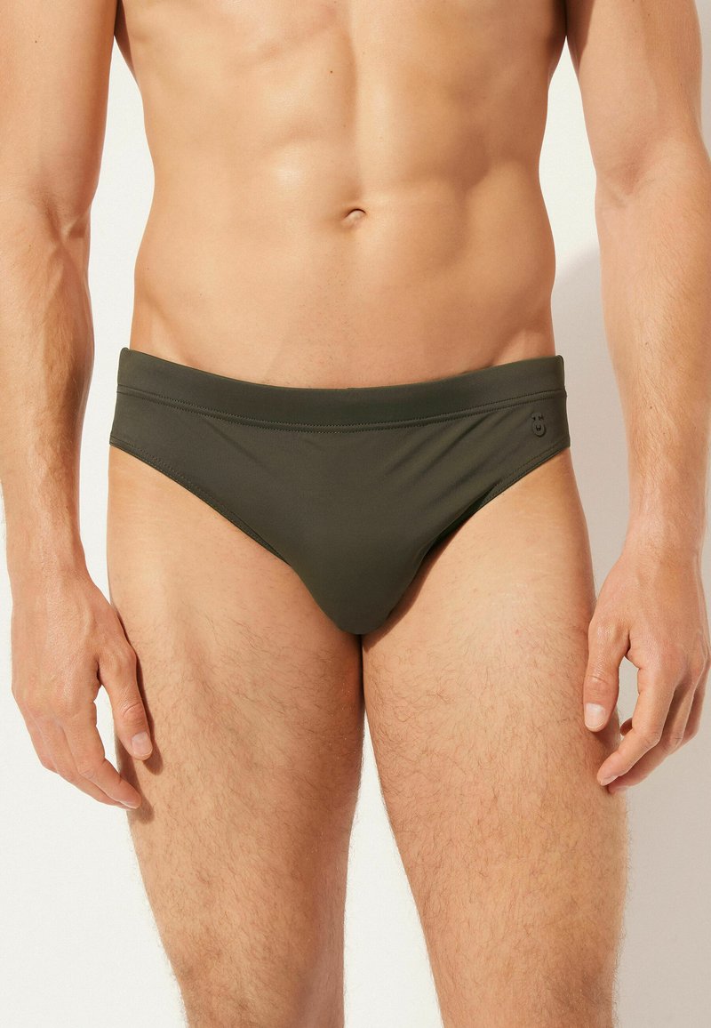 Dunkelgrüne Schwimmbrieftaschen mit glatter Textur, niedriger Taille und breitem Bündchen. Merkmale sind minimale Nähte und ein kleines Logo an der Seite.