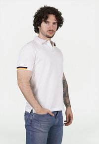 Polo shirt bianco a maniche corte con colletto, caratterizzato da accenti a strisce colorate sulle maniche. Indossato con jeans blu. Tessuto liscio.