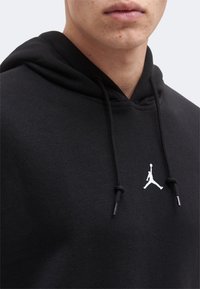 Κοντινό πλάνο ενός ατόμου που φοράει μαύρο φούτερ με κουκούλα με μαύρα κορδόνια και λευκό λογότυπο καλαθοσφαιριστή στη μέση του στήθους.