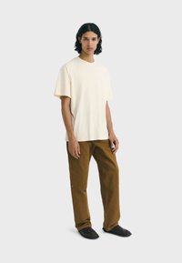 Crème katoenen t-shirt met korte mouwen, gecombineerd met broek met elastische tailleband in bruin en donkere slippers, gedragen door een model dat tegen een effen achtergrond staat.