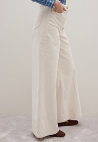 Pantaloni beige a gamba larga realizzati in cotone, con vita alta, ampie tasche laterali e una superficie liscia e texturizzata.