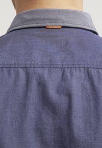 Takapuolet näkymä henkilöstä, joka käyttää sinistä kauluspaitaa, jossa on pieni oranssi "BOSS ORANGE" -labeli niskan takana.