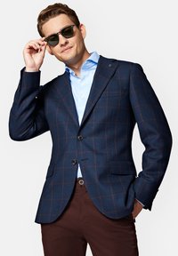 Granatowa marynarka w kratę z brązowymi akcentami, z dwoma guzikami i klapami w kształcie notcha, zestawiona z jasnoniebieską koszulą i ciemnobrązowymi spodniami.