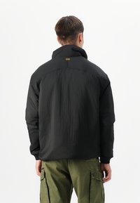 Veste noire avec une surface texturée, col haut et étiquette de logo jaune à l'arrière. Portée avec un pantalon cargo vert olive.