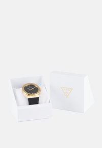 Reloj de pulsera dorado con esfera negra y correa de goma, exhibido en una caja blanca con un logo, que presenta un interior acolchado.