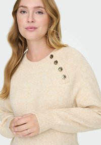Maglione color crema con collo tondo, polsini a coste e cinque bottoni decorativi dorati sulla spalla sinistra. Morbido tessuto a maglia.