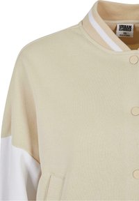 Krémová varsity bunda s béžovým límcem, bílými rukávy, kulatým výstřihem, zapínáním na knoflíky a detaily postranních kapes.