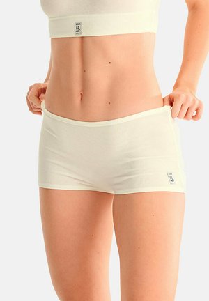 Femme portant un short côtelé crème et un crop top, tirant légèrement vers l’extérieur la ceinture du short avec les deux mains.
