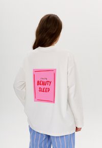 Weißes, übergroßes Sweatshirt mit einem pinken rechteckigen Grafikdruck auf dem Rücken, der den Text "Ich brauche meinen Schönheitsschlaf" in fetten roten Buchstaben zeigt.